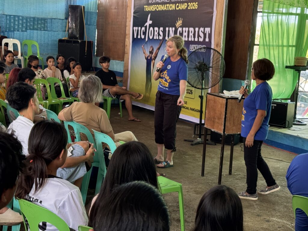 A woman stands on a small stage, speaking into a microphone during a session. She is wearing a blue event t-shirt and black pants. Another woman stands nearby holding a microphone, while a seated audience of young people listens attentively.
