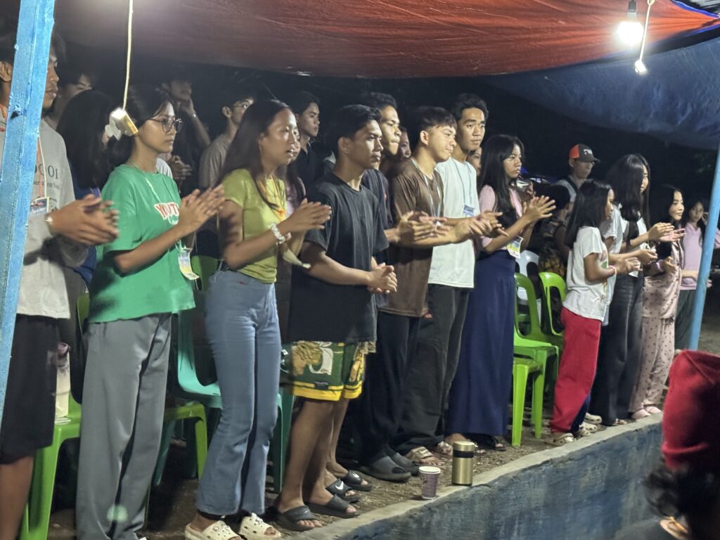 A night-time shot of a row of young people standing and clapping during a worship service. They are outdoors or under a makeshift cover, lit by overhead bulbs, showing a sense of communal energy.