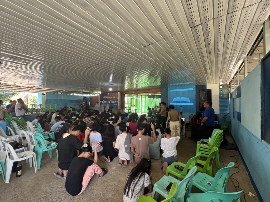 A wide interior shot of a packed hall where dozens of participants are kneeling on the floor in prayer. Many have their hands raised toward a projection of an open Bible on the wall.