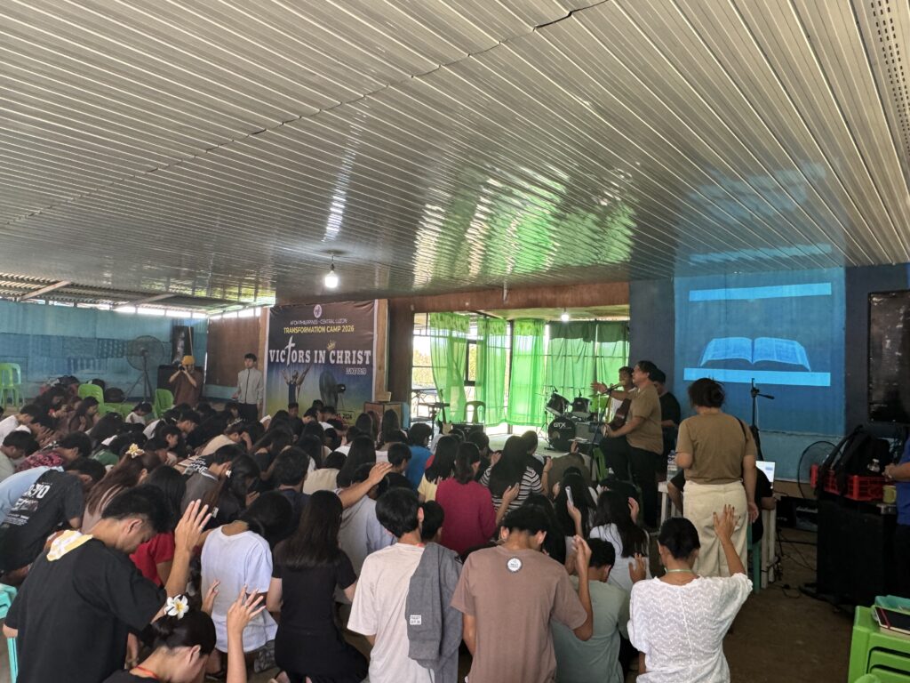 A wide shot from the back of a large group of youth seated in bright green and blue plastic chairs under a large red tarp. They are facing a stage where a presentation is being projected. The atmosphere is casual yet focused.
